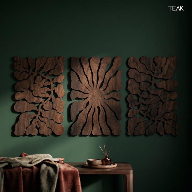 K&ouml;nnte beinhalten: Drei dunkelbraune Holz-Wandpaneele mit floralem Design sind an einer dunkelgr&uuml;nen Wand angebracht. Die Paneele sind aus Teakholz gefertigt. Ein Holztisch mit Schale, Tablett und Diffusor befindet sich im Vordergrund.