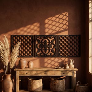 May include: Three dark brown decorative wall panels with intricate geometric and floral patterns. The panels are mounted on a terracotta-colored wall above a wooden console table. The table features two woven baskets, vases, and dried pampas grass.