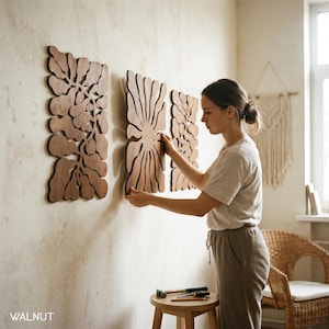 K&ouml;nnte beinhalten: Drei Wandkunst-Paneele aus Walnussholz mit aufwendigen floralen Designs werden an einer hellen Wand ausgestellt. Eine Person ordnet die Paneele an. Das Wort "WALNUT" ist unten links sichtbar.