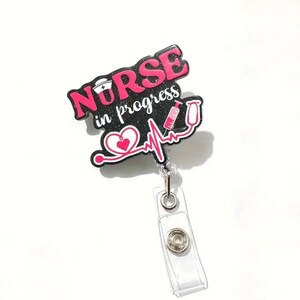 May include: A badge reel with the words "Nurse in progress" in pink and black. The design includes a nurse's hat, a stethoscope, a heart, and a syringe. The reel has a clear plastic clip.