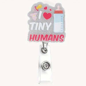 May include: A badge reel with a glittery gray background and the words "I LOVE TINY HUMANS" in white and pink. The design includes a pacifier, a onesie, a heart, and a baby bottle. The reel has a clear plastic strap and a metal clip.