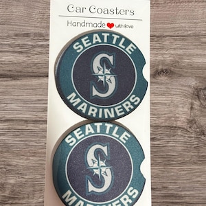 May include: Two round car coasters featuring the Seattle Mariners logo with a teal and navy blue color scheme. The text reads Car Coasters Handmade with love. The coasters are displayed on a white rectangular backing card with a notched edge.