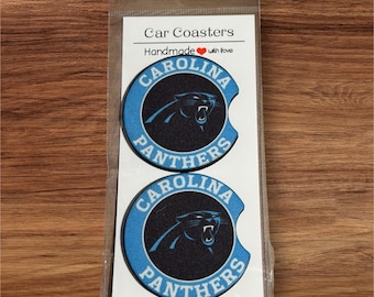 Op de afbeelding: Twee Carolina Panthers auto onderzetters. Elke onderzetter heeft een zwart panterlogo op een blauwe en zwarte achtergrond. De tekst "CAROLINA PANTHERS" is rond het panterlogo gedrukt. De verpakking leest "Car Coasters" en "Handmade with love".