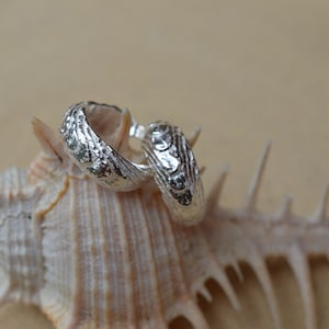 May include: Two silver hoop earrings with a textured surface, resting on a seashell.