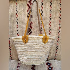 Puede incluir: Bolso tote de paja tejida con asas y detalles de cuero marrón claro. El bolso tiene forma redondeada y color natural, con interior claro. Las asas están sujetas con remaches dorados. El fondo presenta un textil tejido con patrones de zigzag rojos y negros.