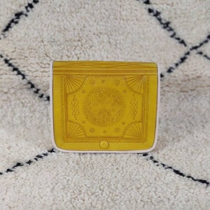 May include: Mustard yellow leather crossbody bag with embossed floral and geometric patterns. The bag has a flap closure with a small button and a light pink interior. The bag is sitting on a white rug with black lines.