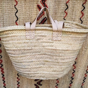 Puede incluir: Un bolso tote de paja tejida con asas y detalles de cuero de color claro. El bolso tiene una parte superior ancha y abierta y una forma redondeada. El fondo es una tela texturizada con patrones de zigzag rojos y negros.