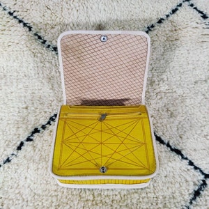 May include: Open yellow handbag with a geometric design on the front. The bag has a white trim and a beige interior with a diamond pattern. The bag is open, revealing a zippered pocket.