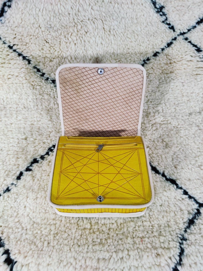 May include: Open yellow handbag with a geometric design on the front. The bag has a white trim and a beige interior with a diamond pattern. The bag is open, revealing a zippered pocket.