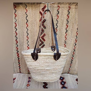 Puede incluir: Bolso tote de paja tejida con asas y detalles de cuero marrón. El bolso tiene forma redondeada y una construcción robusta. Las asas están sujetas con remaches. El fondo presenta un tapiz de pared tejido texturizado.