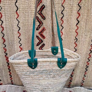 May include: Woven straw tote bag with teal leather handles and accents. The bag has a rounded shape and a natural color, with a textured weave. The handles are attached with silver rivets.