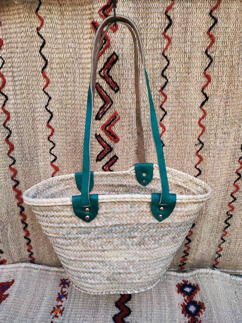 May include: Woven straw tote bag with teal leather handles and accents. The bag has a rounded shape and a natural color, with a textured weave. The handles are attached with silver rivets.