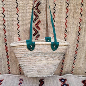 May include: Woven straw tote bag with green leather handles and accents. The bag has a wide, open top and a natural, beige color. The handles are attached with silver rivets. The background features a woven textile with red and black zigzag patterns.