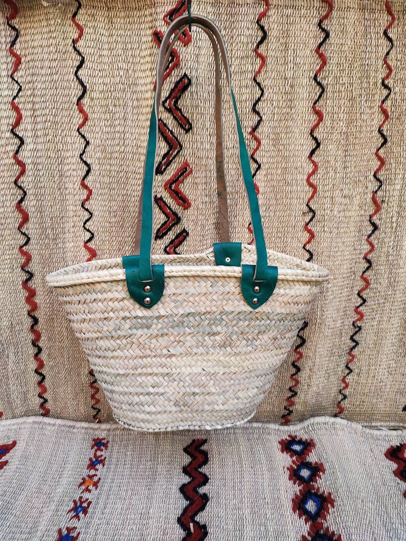 May include: Woven straw tote bag with green leather handles and accents. The bag has a wide, open top and a natural, beige color. The handles are attached with silver rivets. The background features a woven textile with red and black zigzag patterns.