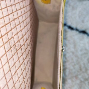 May include: An open, rectangular tote bag with a cream-colored interior and a yellow and cream exterior. The interior features a brown grid pattern. Yellow accents include a small flap with a snap closure and a small handle.