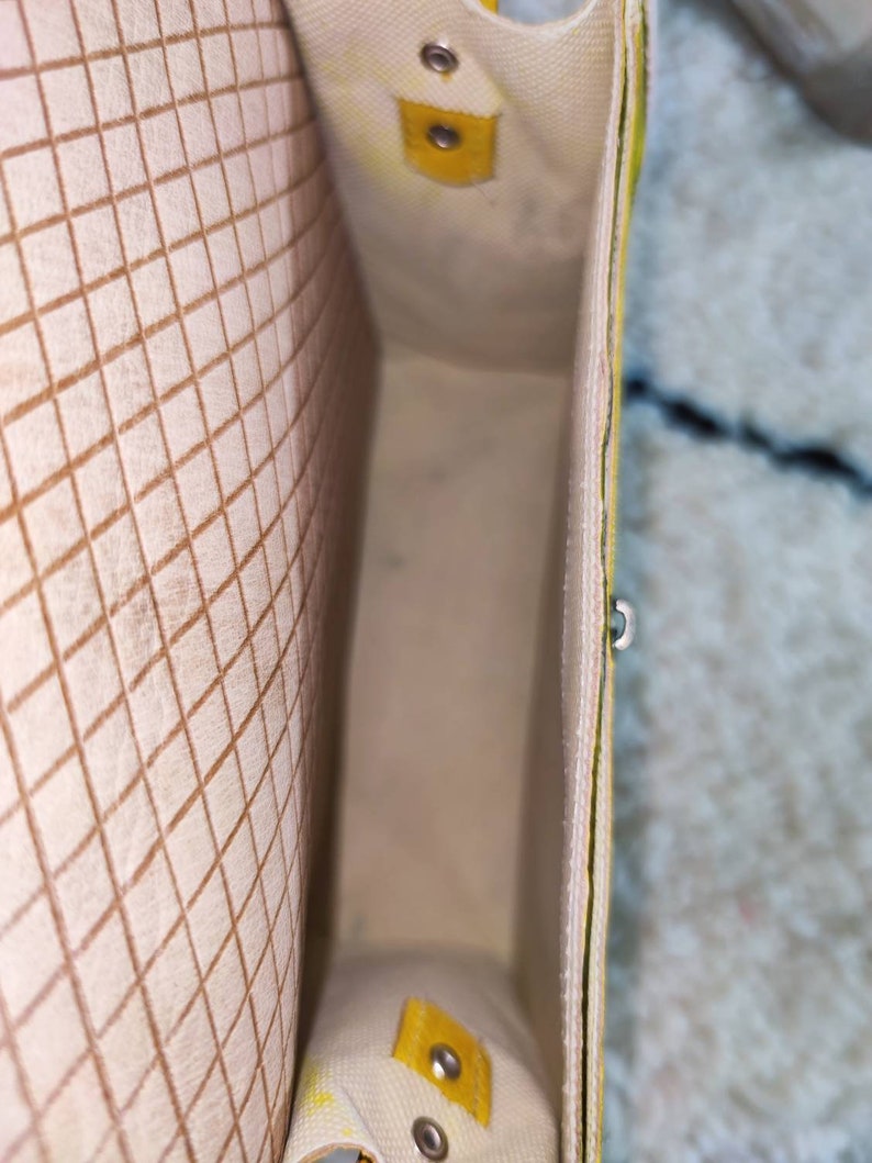 May include: An open, rectangular tote bag with a cream-colored interior and a yellow and cream exterior. The interior features a brown grid pattern. Yellow accents include a small flap with a snap closure and a small handle.