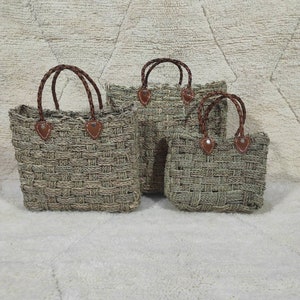 Puede incluir: Tres bolsas de mano tejidas de hierba marina de varios tamaños. Cada bolsa presenta asas trenzadas marrones y detalles de cuero. Las bolsas son de color verde-gris natural y tienen un diseño de tejido de cesta texturizado. Las bolsas se muestran sobre una superficie blanca texturizada.