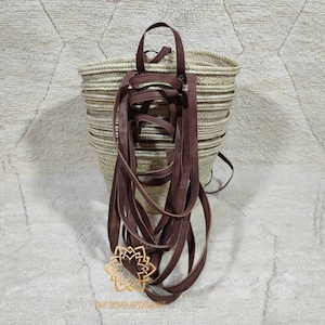 Puede incluir: Una cesta tejida con fibras naturales con un asa de cuero marrón y largas correas de cuero colgantes. La cesta tiene forma redonda. Las correas son de color marrón oscuro. El logotipo de Boho Artshamat es visible.