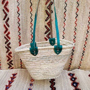 May include: A woven straw tote bag with teal leather handles and accents. The bag is held up against a patterned woven background. The bag's handles are attached with silver rivets.