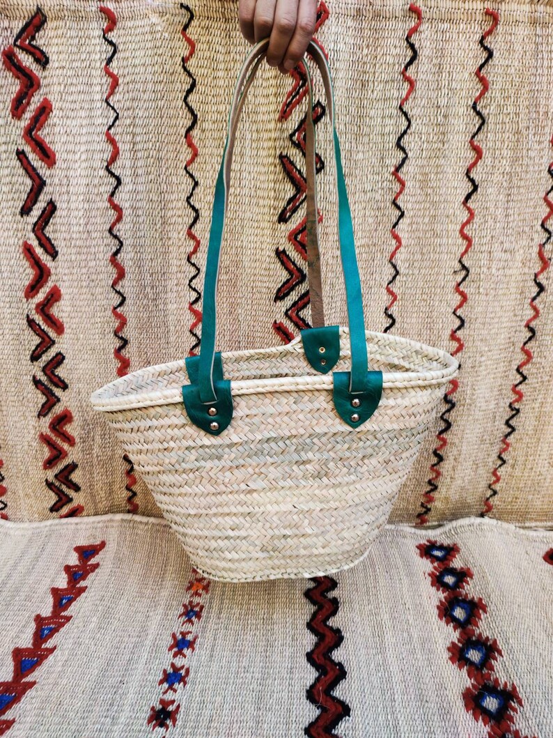 May include: A woven straw tote bag with teal leather handles and accents. The bag is held up against a patterned woven background. The bag's handles are attached with silver rivets.