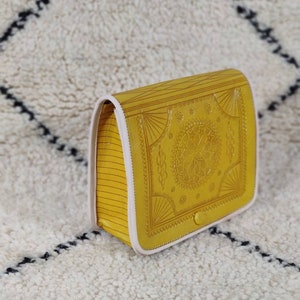 May include: Mustard yellow crossbody bag with a detailed embossed design on the front flap. The bag features a button closure and a white trim. The sides have a striped pattern. The bag is sitting on a white rug with a black diamond pattern.