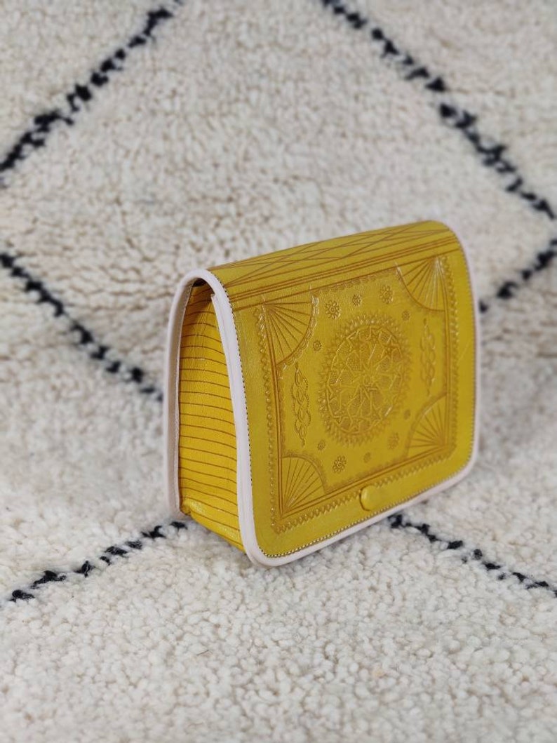 May include: Mustard yellow crossbody bag with a detailed embossed design on the front flap. The bag features a button closure and a white trim. The sides have a striped pattern. The bag is sitting on a white rug with a black diamond pattern.