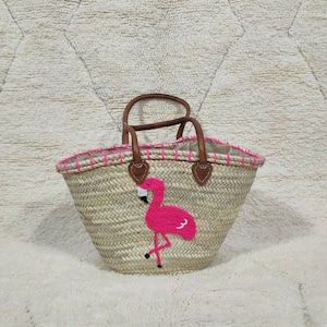Puede incluir: Un bolso tote de paja tejida con un aplique de flamenco rosa. El bolso tiene asas y ribetes de cuero marrón, y costuras rosas a lo largo del borde superior. El flamenco es de color rosa brillante con detalles en negro.