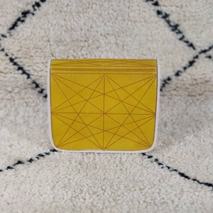 May include: A mustard yellow clutch with a geometric pattern of intersecting lines. The clutch has a white border and a flap closure. The bag is sitting on a white rug with black diamond-shaped patterns.