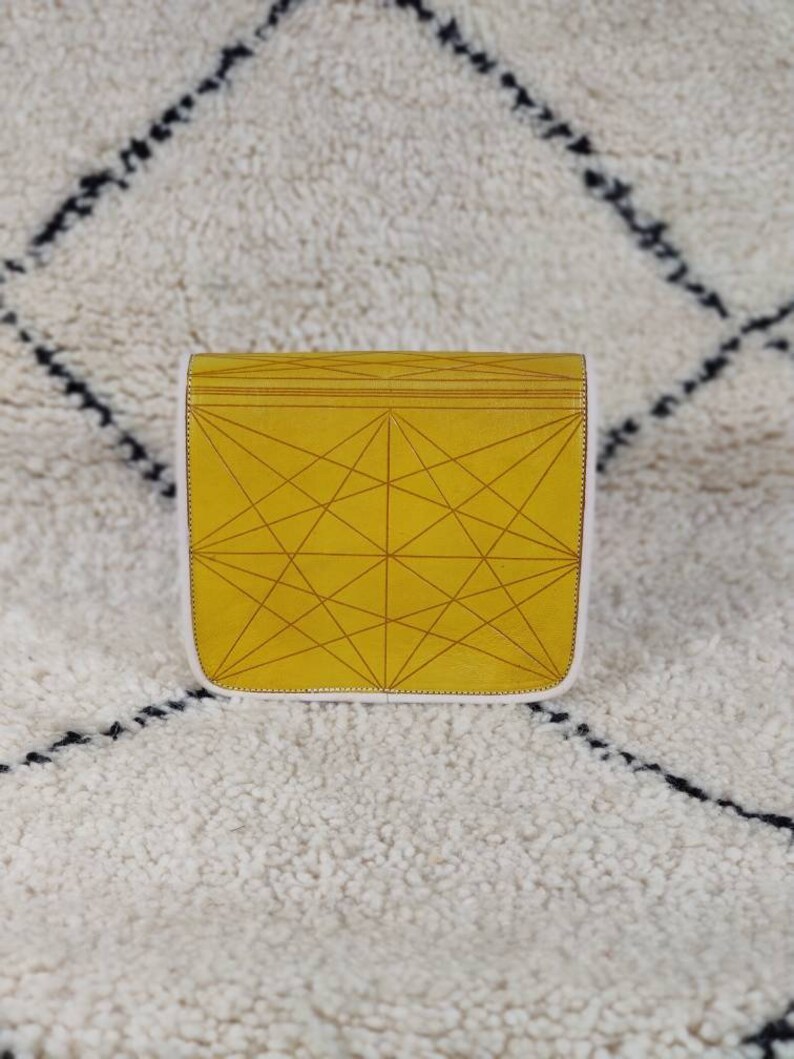 May include: A mustard yellow clutch with a geometric pattern of intersecting lines. The clutch has a white border and a flap closure. The bag is sitting on a white rug with black diamond-shaped patterns.