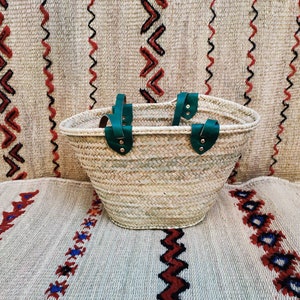 May include: A woven straw tote bag with green leather handles. The bag is oval-shaped and sits on a patterned rug with red, black, and blue geometric designs. The background features a similar rug with vertical patterns.