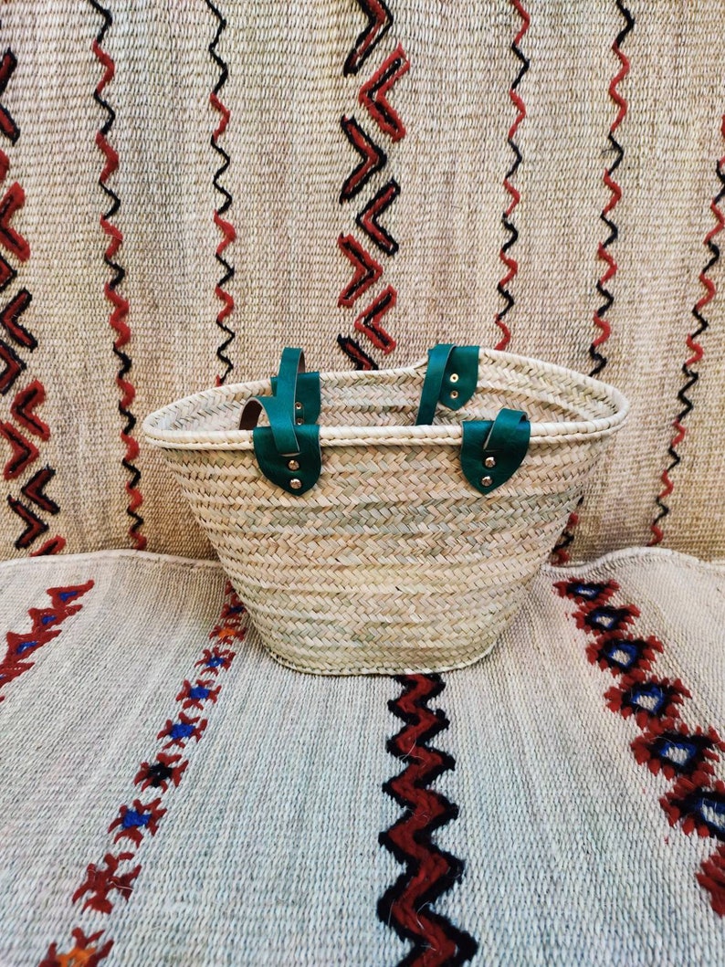 May include: A woven straw tote bag with green leather handles. The bag is oval-shaped and sits on a patterned rug with red, black, and blue geometric designs. The background features a similar rug with vertical patterns.