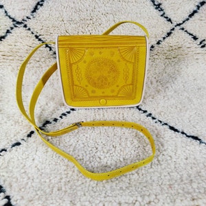 May include: Mustard yellow crossbody bag with a decorative embossed design on the front flap. The bag has a long, adjustable strap and a white interior. The bag is made of leather and has a metal buckle.