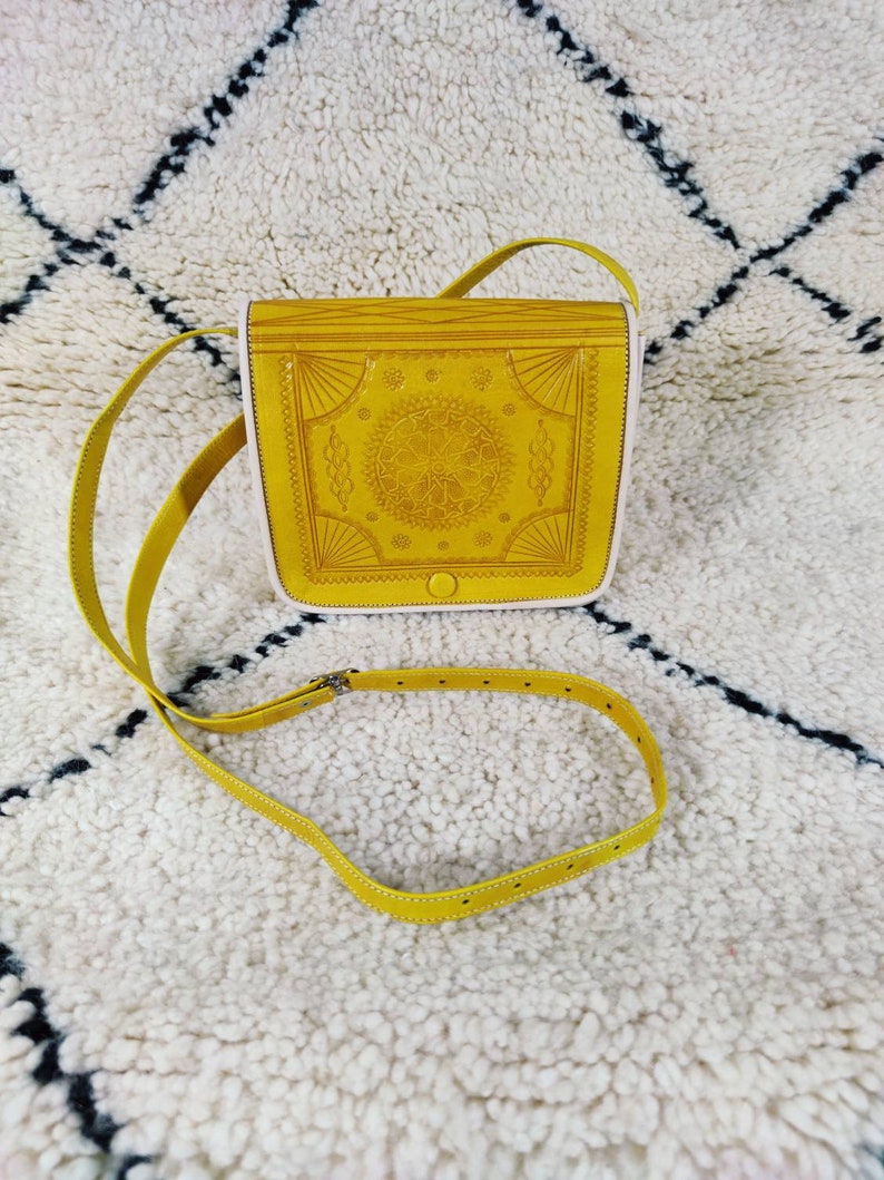 May include: Mustard yellow crossbody bag with a decorative embossed design on the front flap. The bag has a long, adjustable strap and a white interior. The bag is made of leather and has a metal buckle.
