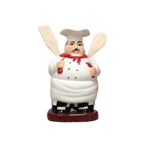 Może przedstawiać: Biała ceramiczna figurka szefa kuchni trzymająca dwa drewniane łyżki. Szef kuchni ma na sobie biały czepek, czerwoną i białą szarfę w paski oraz biały fartuch z czarno-białymi kratami. Szef kuchni stoi na czerwonej podstawie.