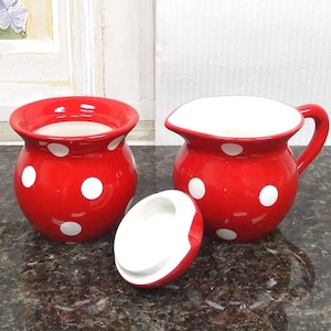May include: Red and white polka dot ceramic creamer and sugar bowl set. The creamer has a handle and the sugar bowl has a lid.