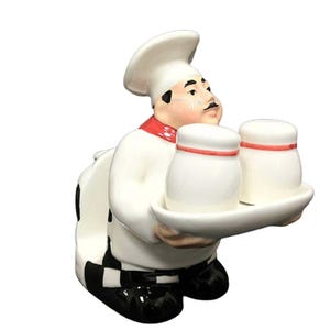 May include: A ceramic chef figurine holding a tray with two white salt and pepper shakers. The chef wears a white uniform with a red neckerchief and a tall chef's hat. The base of the chef's uniform is black and white checkered.