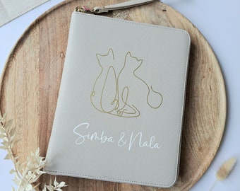 May include: A light gray clutch with a gold zipper and wrist strap. The front features a gold line drawing of two cats and the names "Simba & Nala" in white script. The clutch is on a wooden tray with a white and beige background.