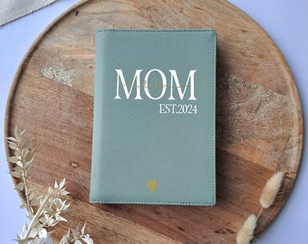 May include: A light green journal with the word "MOM" in white lettering and a gold heart design. The journal also has the text "EST. 2024" in white lettering.