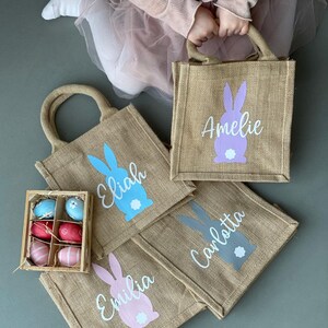 Op de afbeelding: Vier gepersonaliseerde jute paastassen met konijnen silhouetten en namen: Amelia, Elijah, Emilia en Carlotta. De tassen zijn beige met lichtblauwe, roze, paarse en grijze konijnen. De tassen zijn gerangschikt op een grijs oppervlak met een kleine houten doos met kleurrijke paaseieren.