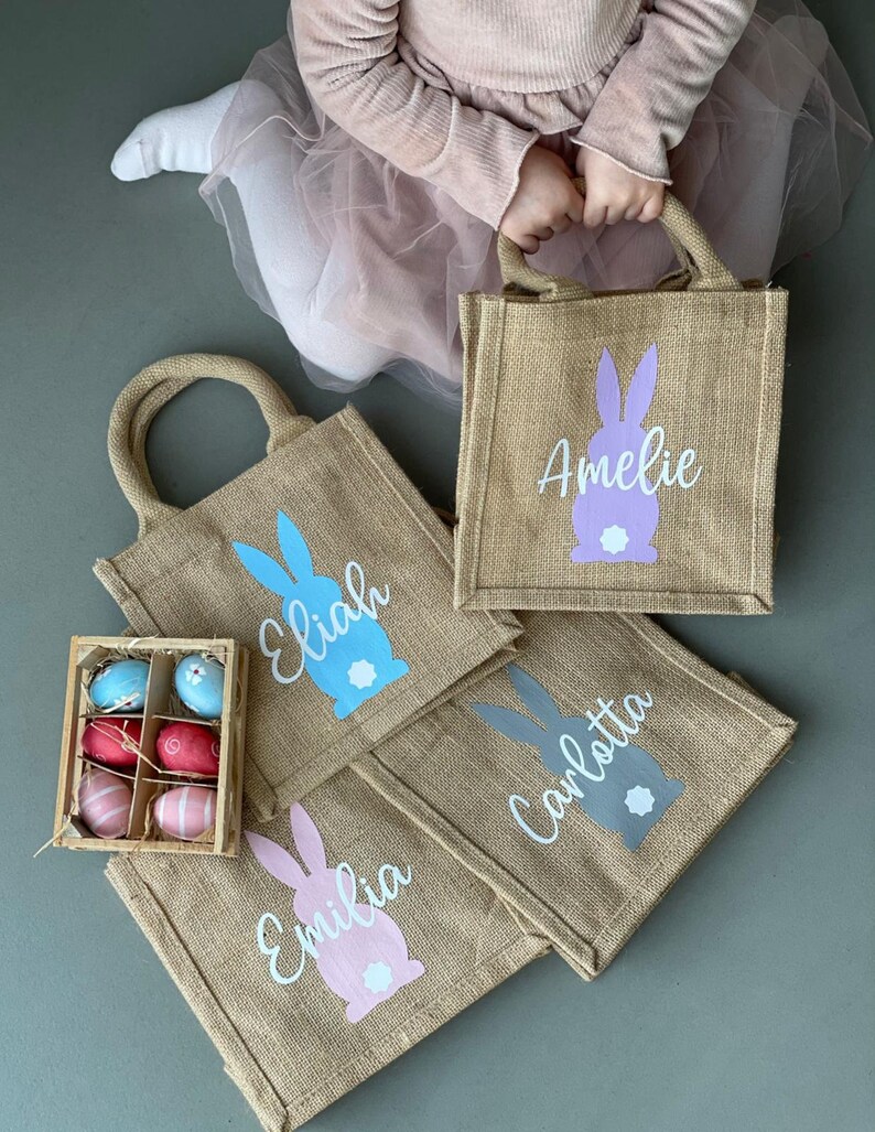 Op de afbeelding: Vier gepersonaliseerde jute paastassen met konijnen silhouetten en namen: Amelia, Elijah, Emilia en Carlotta. De tassen zijn beige met lichtblauwe, roze, paarse en grijze konijnen. De tassen zijn gerangschikt op een grijs oppervlak met een kleine houten doos met kleurrijke paaseieren.