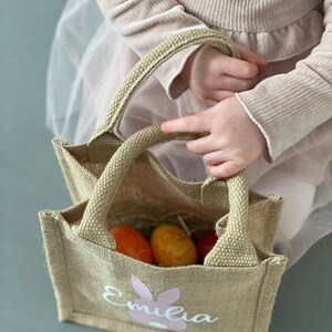Op de afbeelding: Een bruine jute tas met een roze konijn en de naam "Emilia" erop gedrukt. De tas is gevuld met oranje paaseieren.