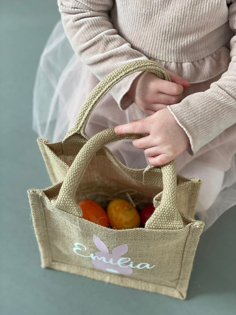 Op de afbeelding: Een bruine jute tas met een roze konijn en de naam "Emilia" erop gedrukt. De tas is gevuld met oranje paaseieren.