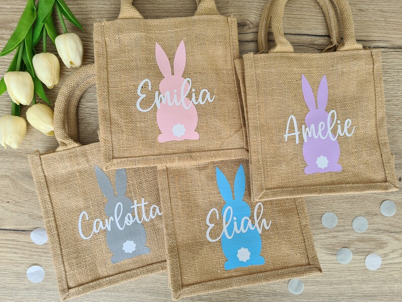 Op de afbeelding: Vier kleine jute tassen met konijnenontwerpen en gepersonaliseerde namen. De tassen zijn in roze, paars, grijs en blauw. De namen op de tassen zijn Emilia, Amelie, Carlotta en Elijah.