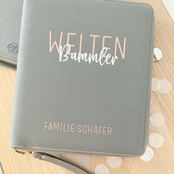 May include: A gray zippered clutch with the words "WELTEN BUMMLER" and "FAMILIE SCHÄFER" in pink lettering. The clutch has a wrist strap and a gold-colored zipper. The background is a light-colored wooden surface.