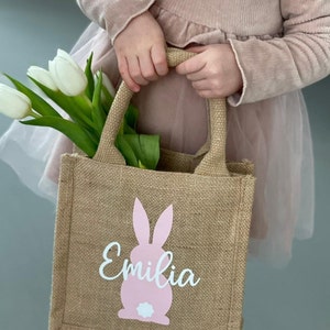 Op de afbeelding: Een bruine jute draagtas met een roze konijn en de naam "Emilia" in witte letters. De tas heeft twee handvatten en wordt vastgehouden door een kind.