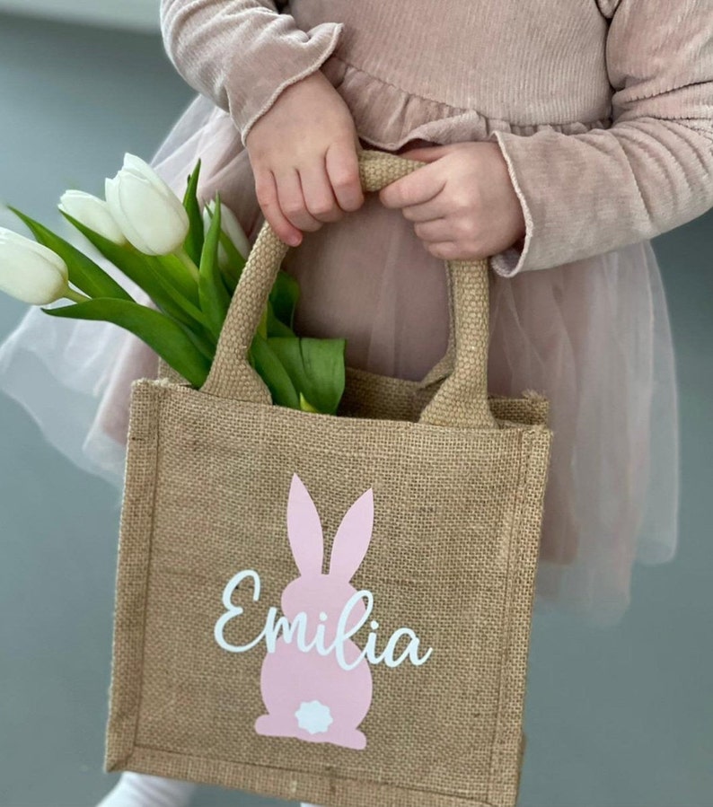Op de afbeelding: Een bruine jute draagtas met een roze konijn en de naam "Emilia" in witte letters. De tas heeft twee handvatten en wordt vastgehouden door een kind.