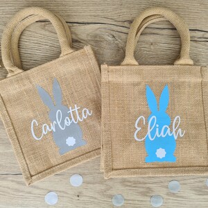 Op de afbeelding: Twee jute tassen met konijnen silhouetten en gepersonaliseerde namen. De tas links heeft een grijze konijn en de naam "Carlotta" in wit. De tas rechts heeft een blauwe konijn en de naam "Elijah" in wit.