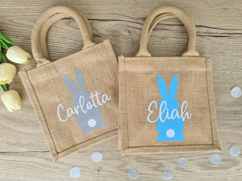 Op de afbeelding: Twee jute tassen met konijnen silhouetten en gepersonaliseerde namen. De tas links heeft een grijze konijn en de naam "Carlotta" in wit. De tas rechts heeft een blauwe konijn en de naam "Elijah" in wit.
