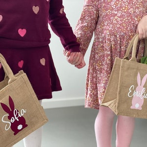 Op de afbeelding: Twee kinderen die gepersonaliseerde jute paasmanden vasthouden met konijnen silhouetten en hun namen, Sofia en Emilia, in witte letters.