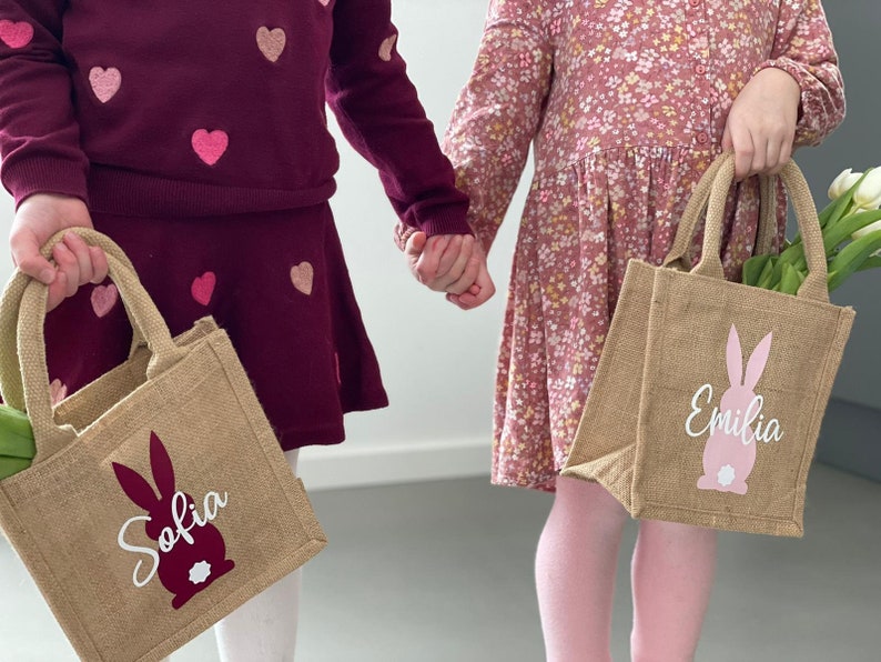 Op de afbeelding: Twee kinderen die gepersonaliseerde jute paasmanden vasthouden met konijnen silhouetten en hun namen, Sofia en Emilia, in witte letters.